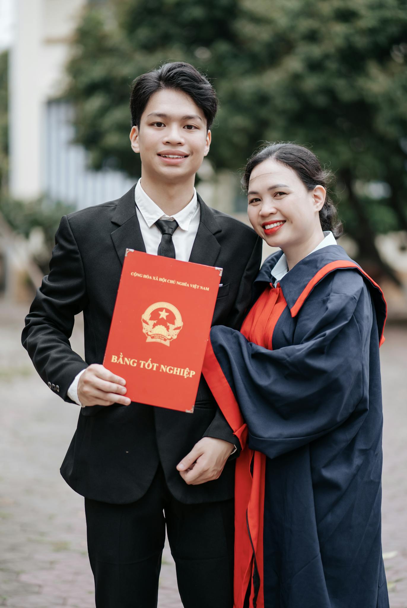Two graduates in formal attire holding a diploma, celebrating their achievement outdoors.